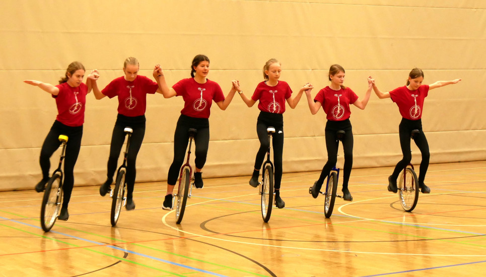 Foto des 6er-Einradfahrens der Junioren U19 der Sportgemeinschaft RSV Kissing/TSV Merching, wobei alle Sportlerinnen nebeneinander in einer Reihe fahren und sich gegenseitig an den Händen halten.